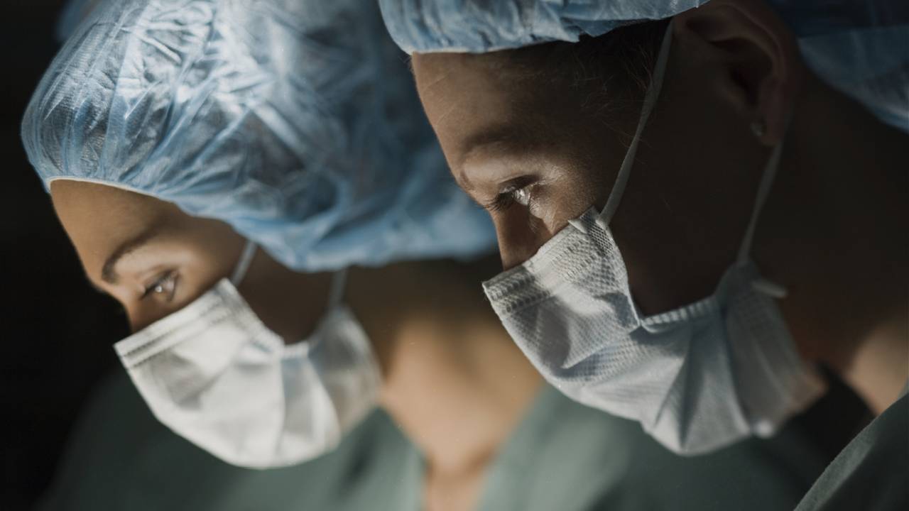 A imagem mostra o rosto de duas mulheres de perfil. Elas estão de máscara e touca no cabelo, em uma sala de cirurgia
