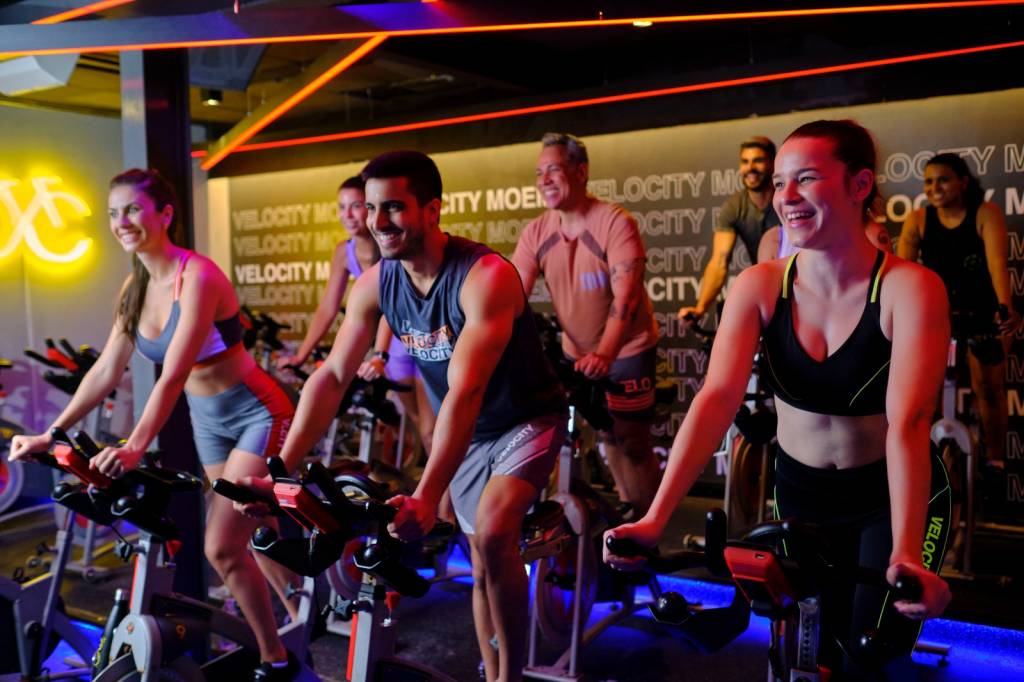 A imagem mostra um grupo de pessoas participando de uma aula de spinning. Elas estão pedalando em bicicletas estacionárias em uma sala de ginástica. O ambiente é iluminado com luzes de neon, com um símbolo de bicicleta amarelo na parede ao fundo.