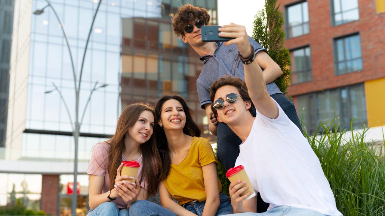 A imagem mostra quatro jovens ao ar livre, tirando uma selfie. Eles estão sorrindo e parecem felizes. Três deles estão sentados em um degrau, enquanto o quarto jovem, de pé atrás, segura o celular para capturar a foto. Duas das pessoas estão segurando copos de café. Ao fundo, é possível ver um prédio moderno de vidro.