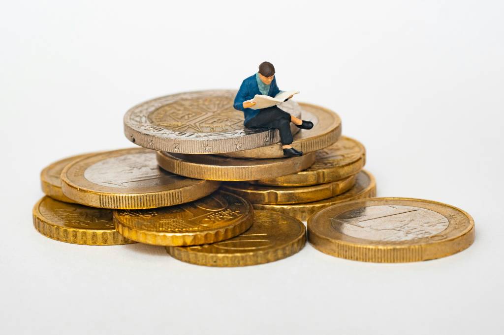 A imagem mostra uma figura em miniatura de uma pessoa sentada em cima de uma pilha de moedas douradas, lendo um jornal. A figura está vestida com roupas formais, e a cena é capturada em um fundo branco, destacando o contraste entre o pequeno tamanho da figura e as moedas maiores.