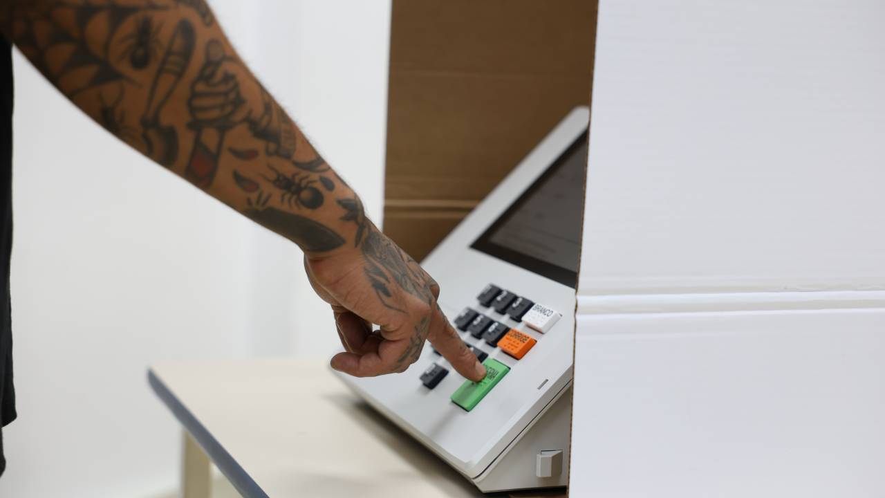 A imagem mostra o braço de uma pessoa tatuada prestes a apertar o botão verde de uma urna eletrônica. A mão está dentro de uma cabine de votação, e o ambiente parece ser um local de eleição.