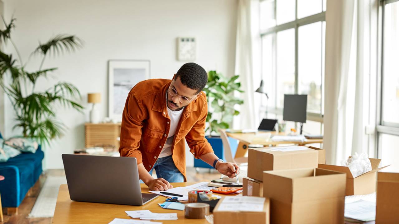 Homem analisando documentos olhando para o notebook em home office.