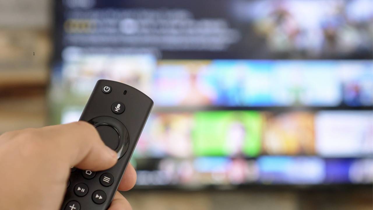 Uma mão segurando um controle remoto. Ao fundo, uma televisão desfocada.